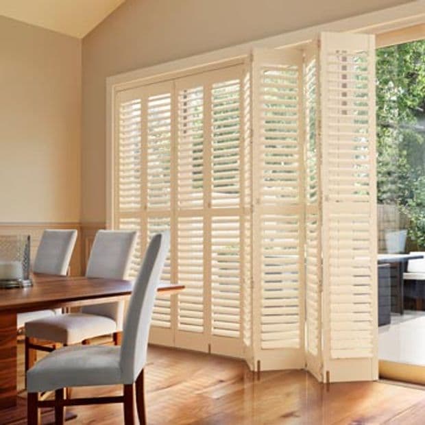 White plantation shutters covering patio doors, several panels folded open at right revealing glass; slatted louvers partly angled to filter sunlight â€” dining room with wooden table, upholstered chairs, garden view.