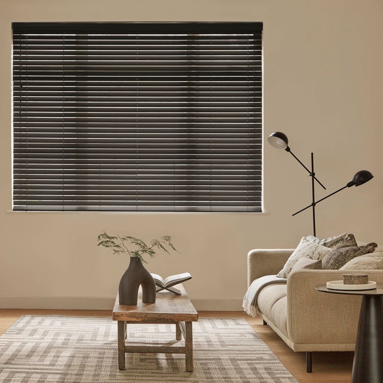Horizontal dark venetian blinds covering a window; fully lowered and closed, admitting thin light bands; above a neutral living room with beige sofa, wooden coffee table and floor lamp.