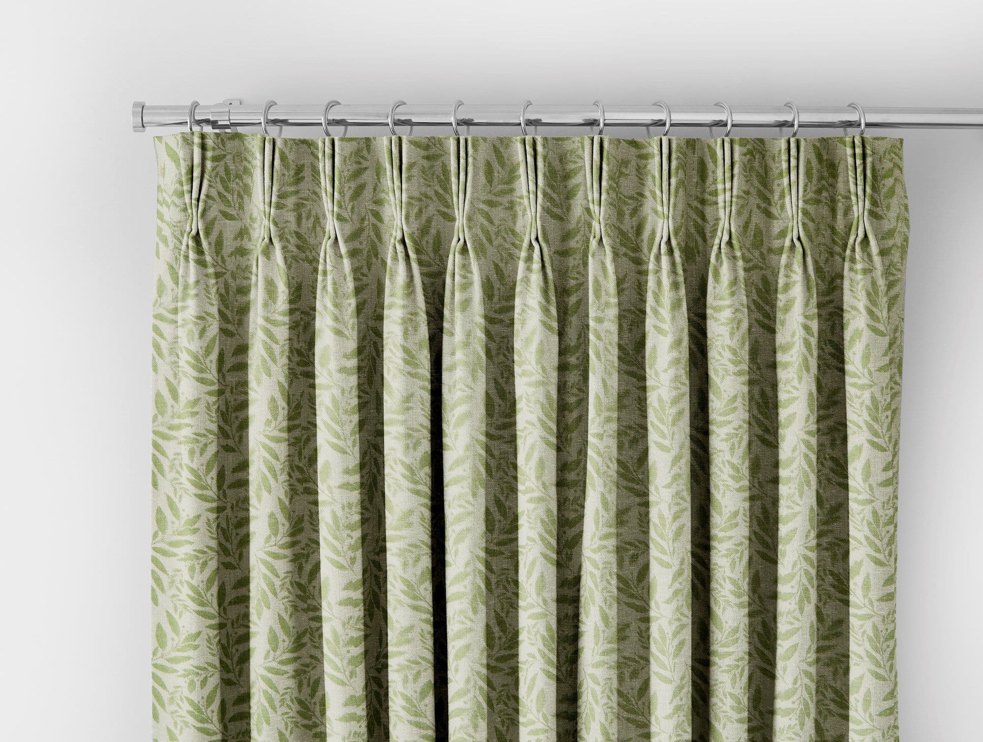 Leaf-patterned green pinch-pleat curtains hung from a chrome rod with ring hooks, neatly closed and pleated, set against a white wall under soft, diffuse lighting.