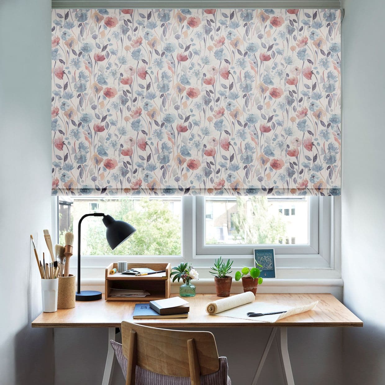 Floral roller blind (pastel watercolor flowers) lowered over a double window, filtering bright daylight; small wooden desk beneath with lamp, potted plants, notebooks, and a view of trees and buildings.