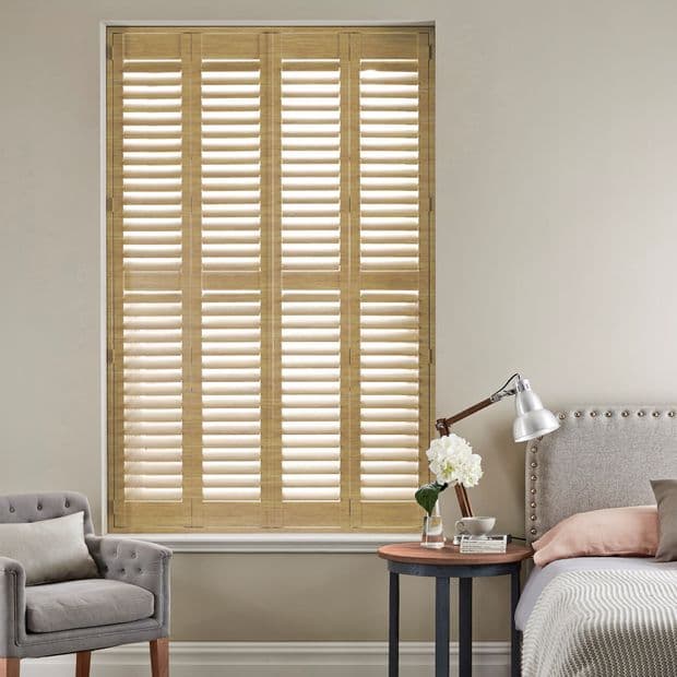Wooden full-height plantation shutters covering tall window, divided into four louvered panels with tilted slats; closed, they softly filter daylight into a neutral bedroom with bed, side table, armchair.