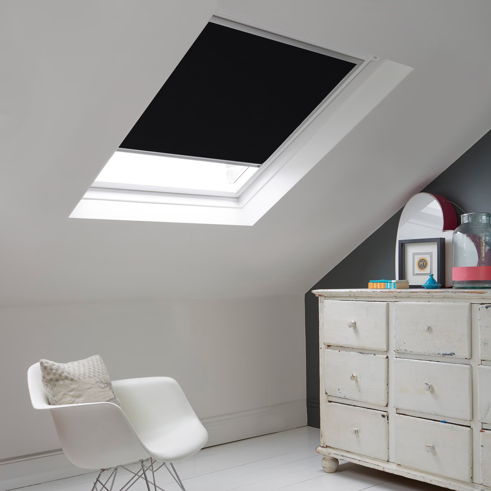 Room with white eaves wall, dark blue-grey wall to right with a shabby chic white drawer unit, a contemporary white chair, white painted floorboards and a skylight blind in black.