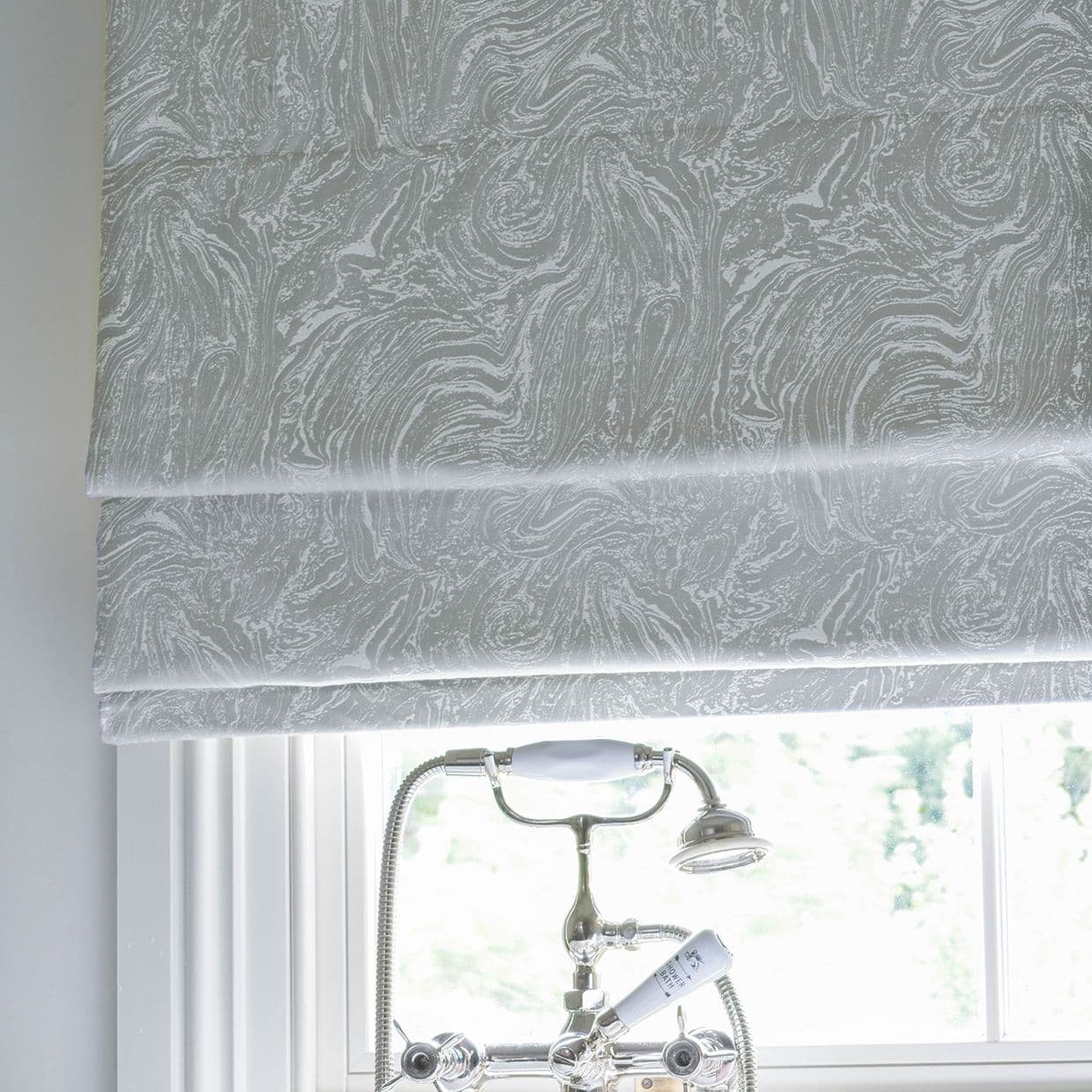 Roman shade with pale gray marbled pattern, lowered in neat horizontal folds, filtering bright daylight over a white-trim window; chrome bathtub faucet and handheld shower visible below in a bathroom.