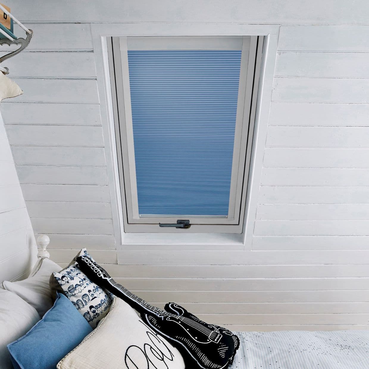 Blue pleated blind over a rectangular skylight, fully lowered and filtering soft daylight; set in white painted tongue-and-groove ceiling above a bed with blue-and-white pillows and a guitar cushion.