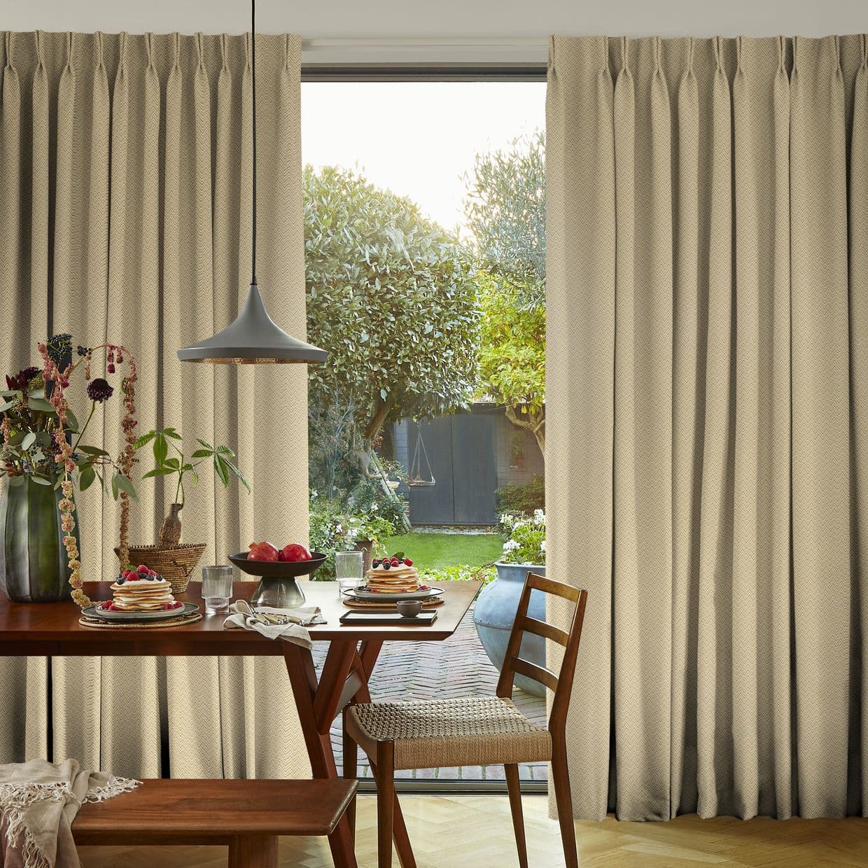 Beige full-length pleated curtains hung at a wide patio window, parted to reveal a sunlit garden; they frame and filter light over a wooden dining table set for breakfast.
