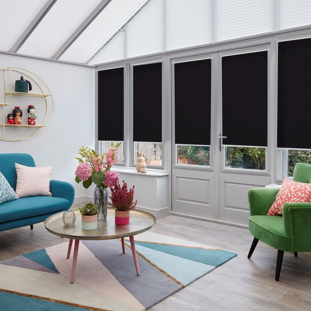 Black roller blinds, lowered across four tall window/door panels with white frames, fully closed over lower panes; blocking light and view while skylight remains bright, in a colorful conservatory lounge.