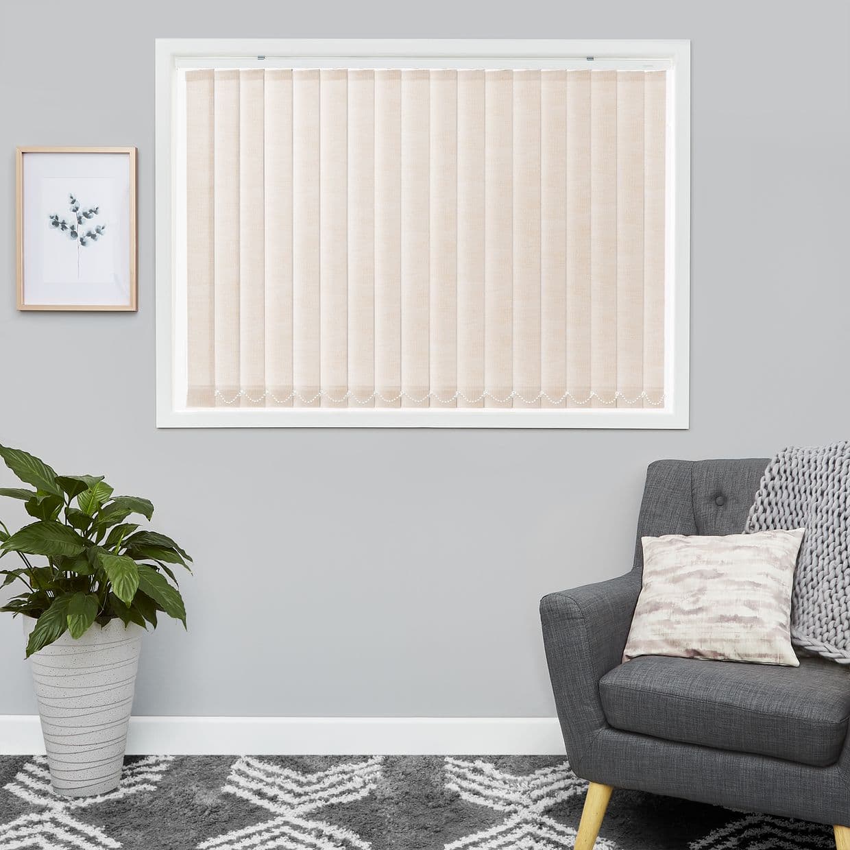 Vertical beige fabric vertical blinds covering the window, closed and filtering soft daylight; set on a gray wall in a living room with framed botanical print, potted plant, gray armchair and patterned rug.