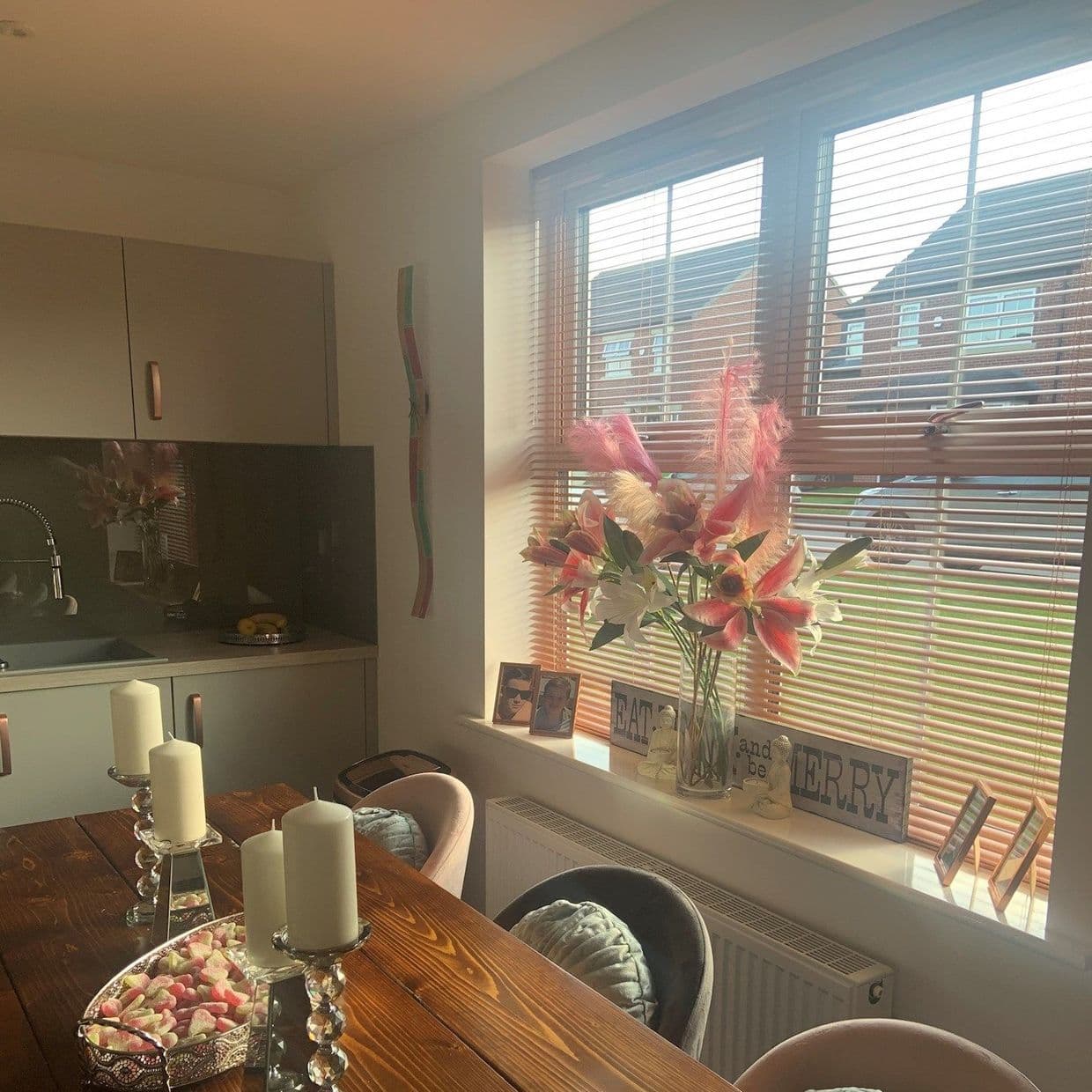 Wooden horizontal blinds over a window, slats tilted to filter warm sunlight; vase of pink lilies on the sill, wooden dining table with candles in a cozy kitchen-diner.

EAT and be MERRY