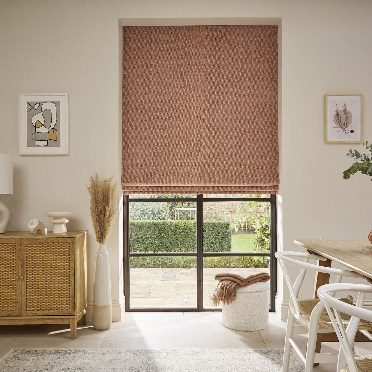 A woven terracotta Roman blind, lowered to mid-window with fold lines, filtering soft daylight over a black-framed patio window in a neutral dining/living area with wooden furniture and garden view.