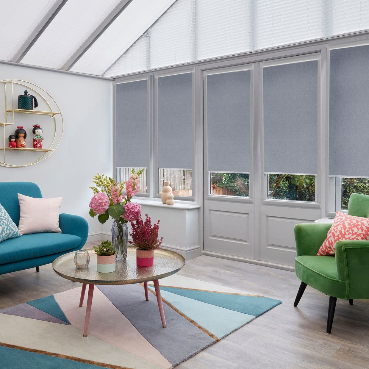 Grey roller blinds, fully lowered over tall doors and windows with narrow bottom gaps, filtering daylight; set in a bright conservatory-style living room with colorful sofas, coffee table and plants.