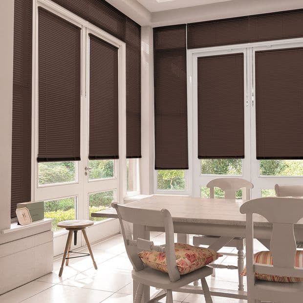 Dark brown pleated blinds covering tall white-framed windows, lowered to block and softly filter daylight; small dining area with white table and chairs, floral cushions and garden view.