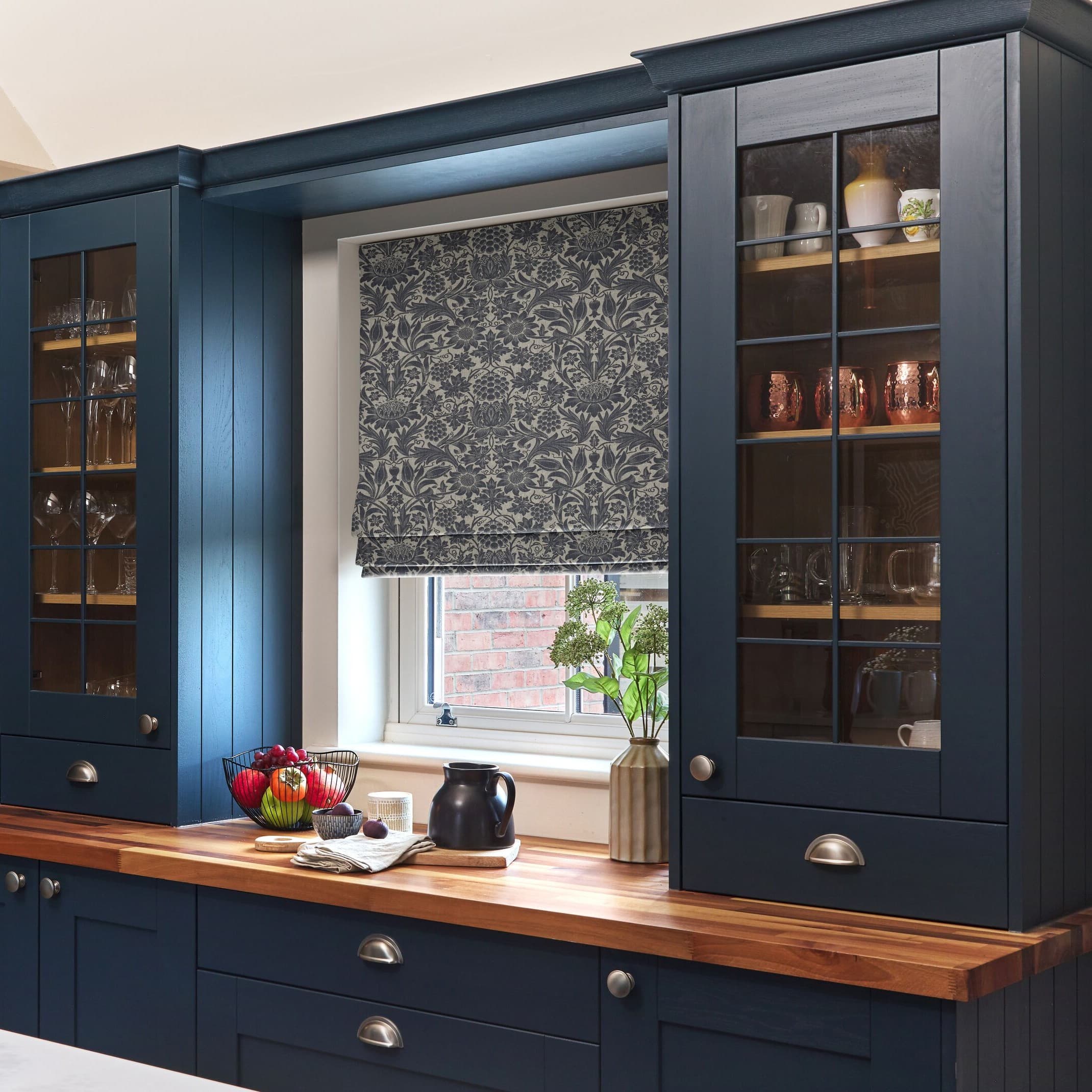 Roman fabric blind with floral pattern, lowered to cover most of the window and filter light; set within navy kitchen cabinetry above a wooden countertop with vase and fruit bowl.