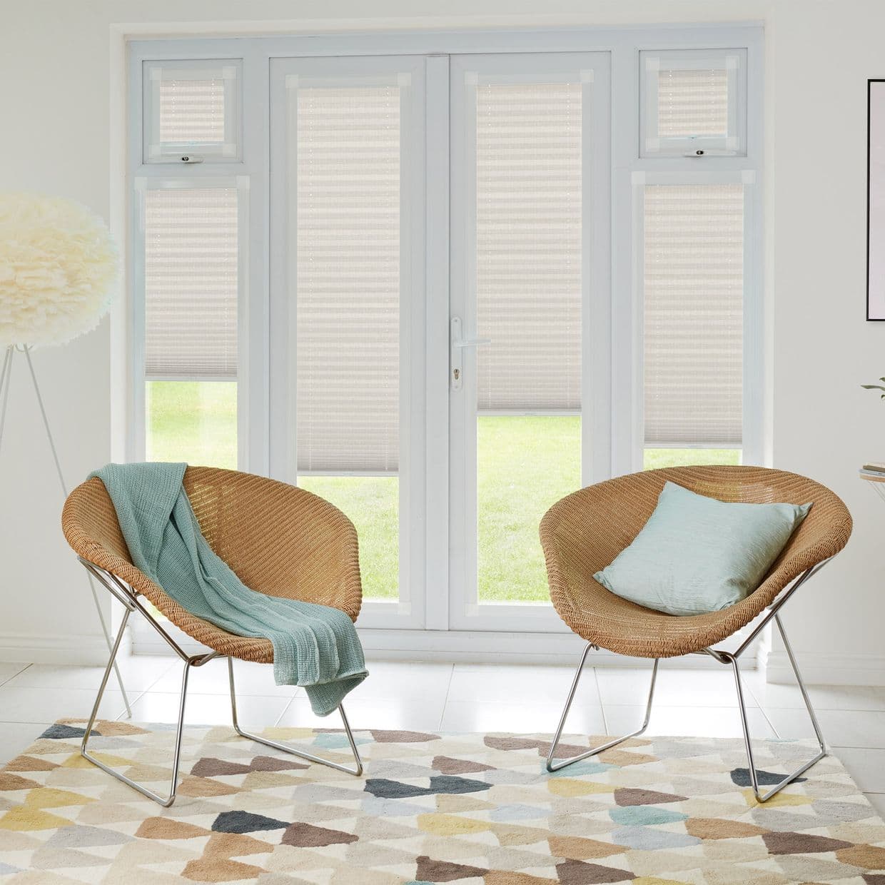 Light beige pleated blinds on French doors and sidelights, lowered midway, filtering soft daylight; two rattan lounge chairs with blue textiles face the doors in a bright, minimalist living room.