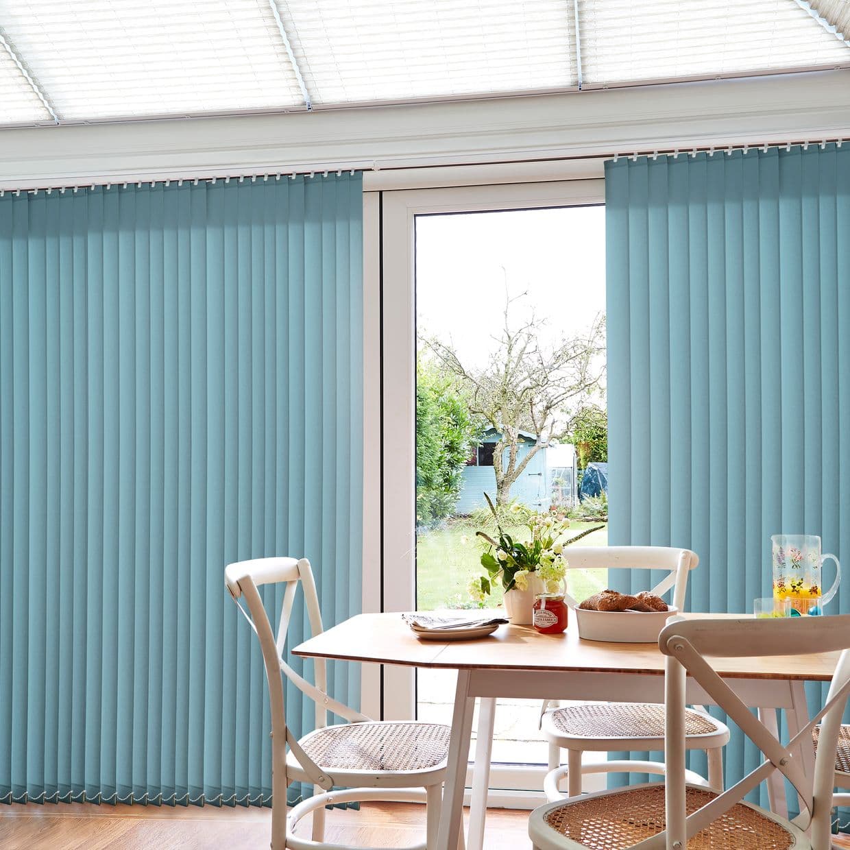 Vertical blue fabric blinds, ceiling-mounted with slats, are parted to reveal a central glass patio door, allowing daylight over a dining table with white chairs and a garden view.