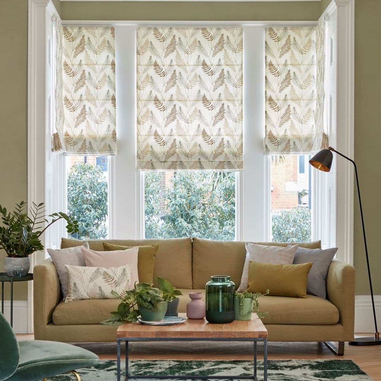 Three patterned roman shades (fern print) covering a threeâ€‘part bay window, partially lowered and filtering daylight; in a living room with beige sofa, cushions, coffee table, plants and floor lamp.