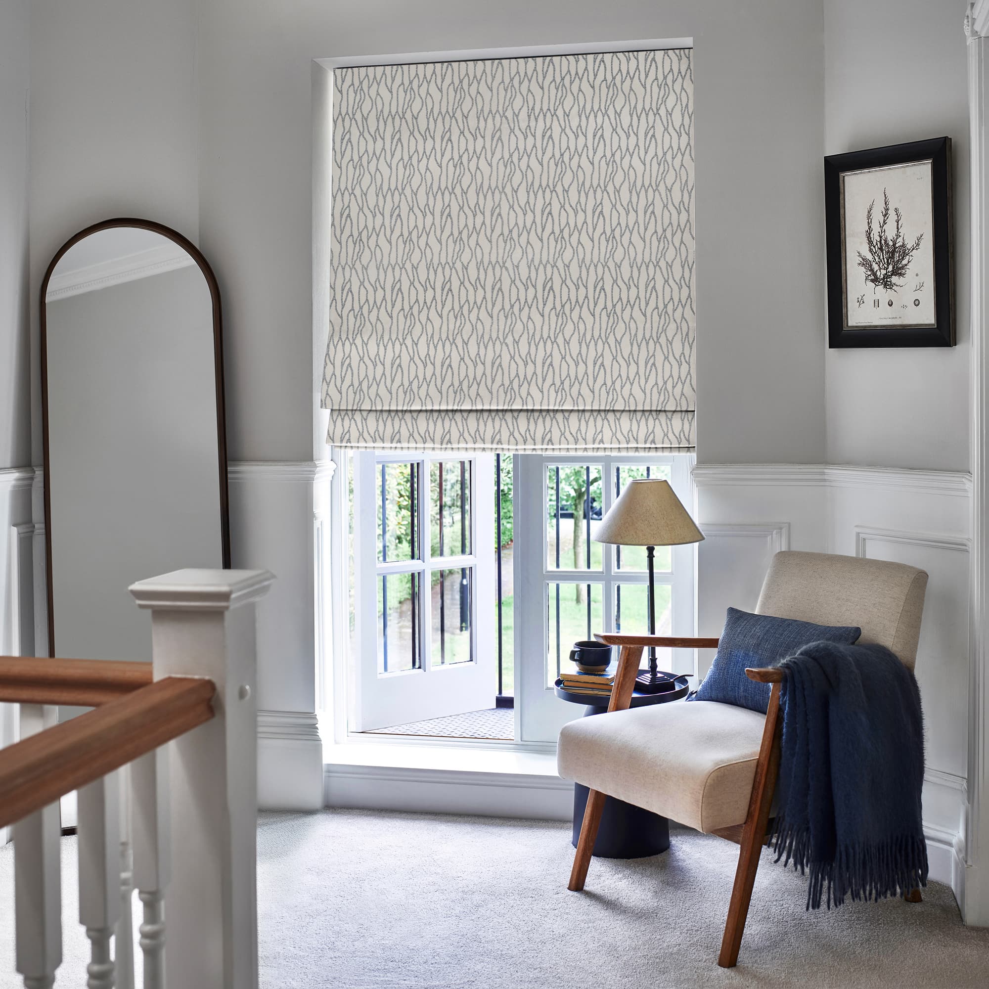 Patterned fabric Roman blind lowered over recessed window, subtle gray wave motif; filtering daylight into a bright upstairs landing with upholstered armchair, navy throw, floor lamp, and garden view.