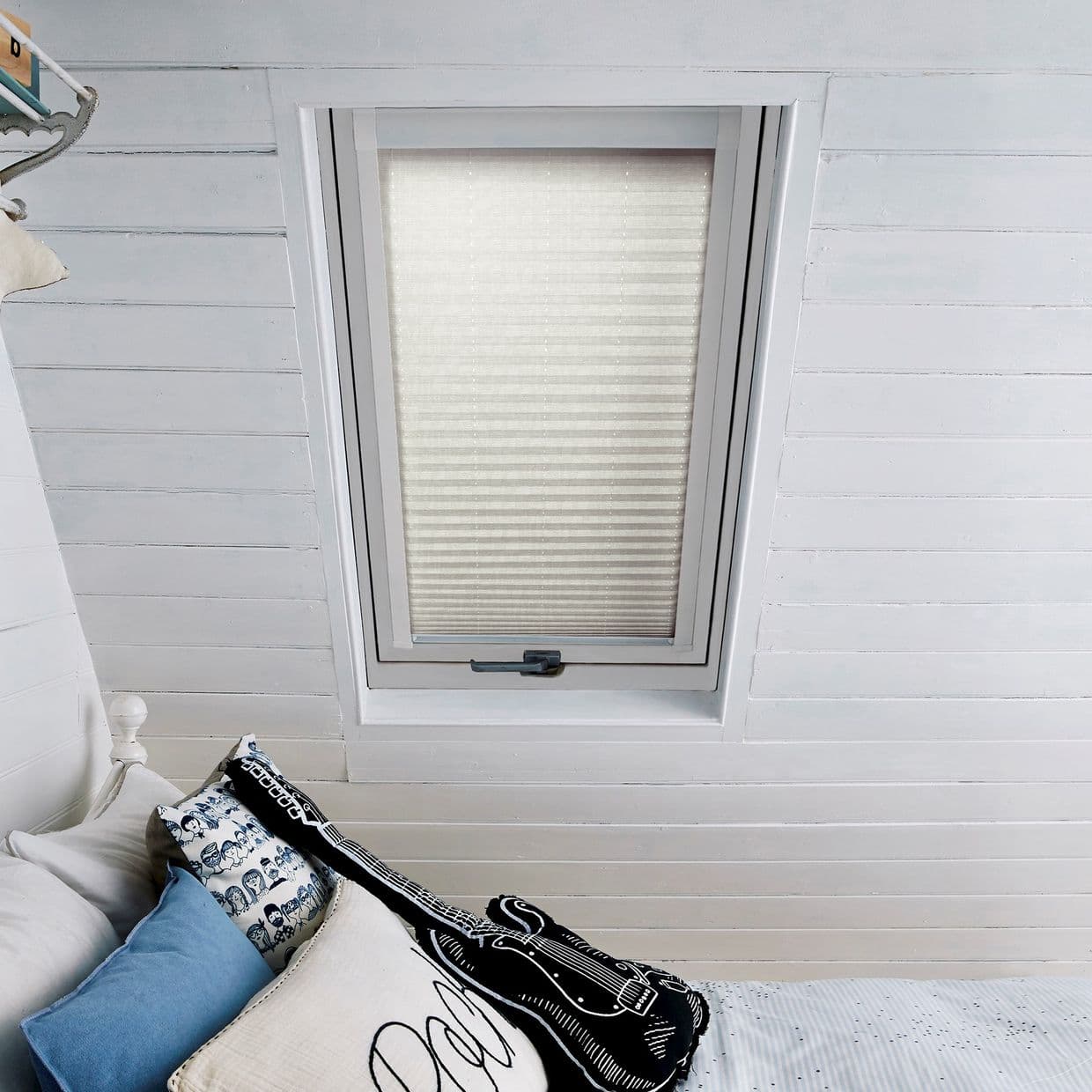 A cream pleated cellular blind, fully lowered over a rectangular skylight, softly diffusing daylight. Set in a white-painted shiplap attic ceiling above a bed with patterned pillows.