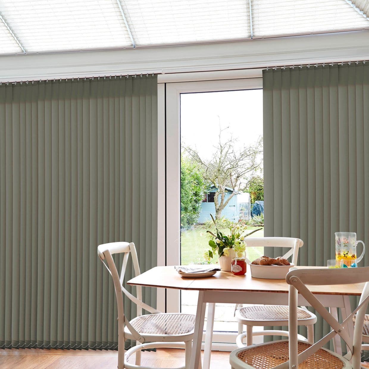 Vertical, floor-length vertical blinds on a sliding glass door; mostly closed with a central gap letting daylight through. Dining table and chairs in a bright conservatory opening onto a backyard garden.