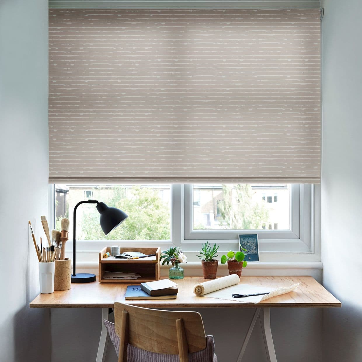 Beige patterned roller blind with thin white horizontal stripes, lowered over the window, filtering daylight; small home workspace with wooden desk, lamp, brushes, potted plants and view of trees.