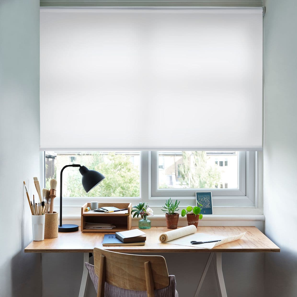 White fabric roller blind, fully lowered across a double window, diffusing daylight; it covers the panes and overlooks a leafy view above a wooden desk with lamp, plants and art supplies.