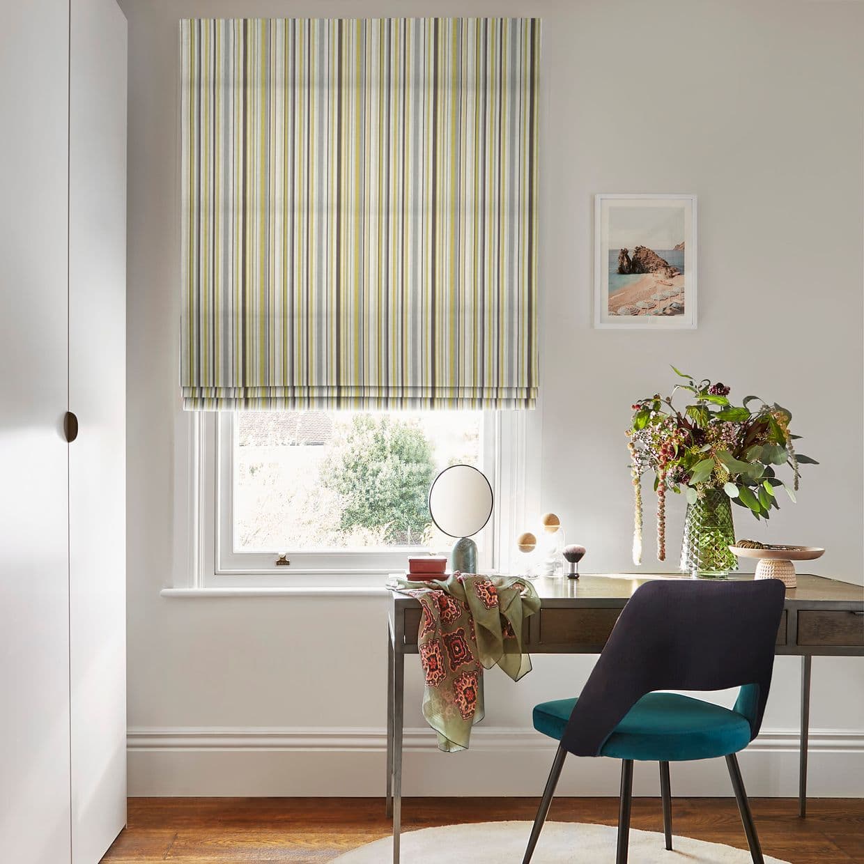 A vertically striped Roman blind (yellow, grey, white) lowered over a sash window, filtering soft daylight into a neutral home office/vanity with desk, teal chair, vase of flowers and mirror.