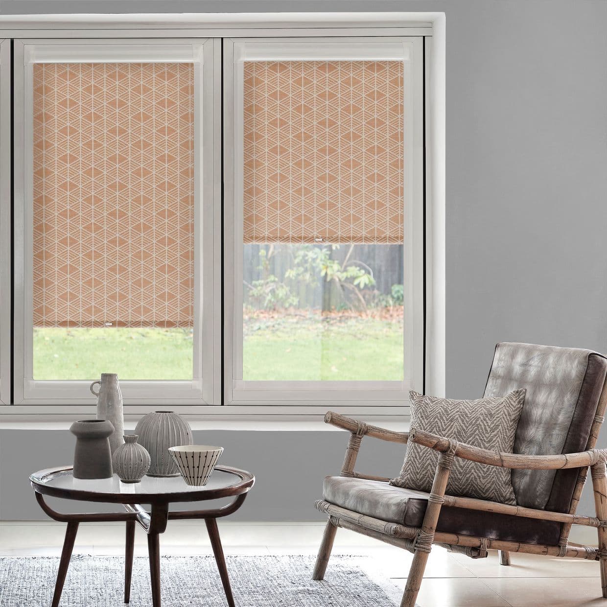 Peach geometric roller blinds across a two-panel windowâ€”left fully lowered, right partially loweredâ€”filtering daylight and revealing a lower yard view; gray-walled living room with wicker armchair and ceramic-decorated coffee table.
