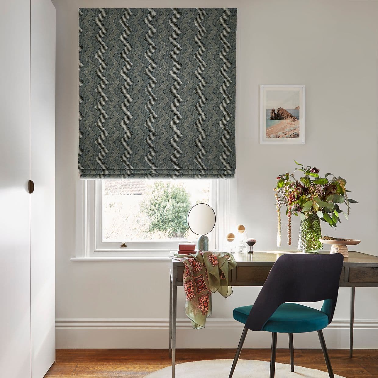 Teal-green chevron Roman blind lowered over the window, mostly closed and filtering daylight. Below, a desk with chair, vase of flowers and framed seaside print in a home office.