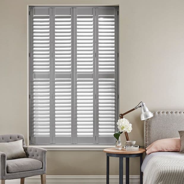 White plantation-style shutters covering a tall window, fully closed with slats slightly tilted to admit soft light; bedroom setting with upholstered bed, side table lamp, armchair, neutral walls.