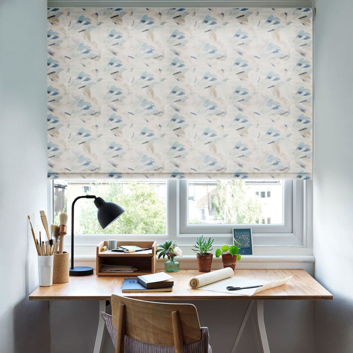 Patterned fabric roller blind, lowered across the window with pale blue-and-beige brushstroke print, diffusing daylight over a wooden desk with lamp, potted plants, notebooks and a suburban view.