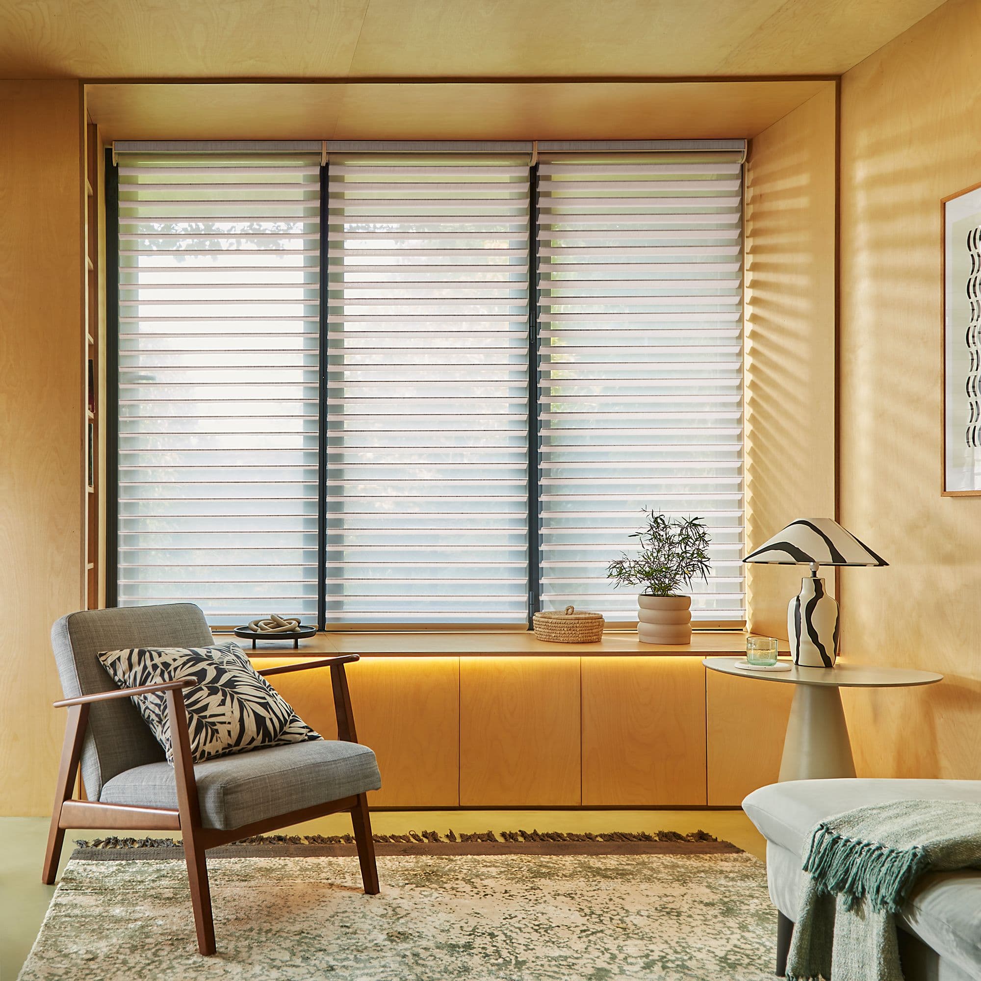 Three-panel horizontal slatted blinds, lowered across a recessed window, filtering soft daylight and casting striped shadows in a warm wood-paneled sitting room with armchair, side table and plant.