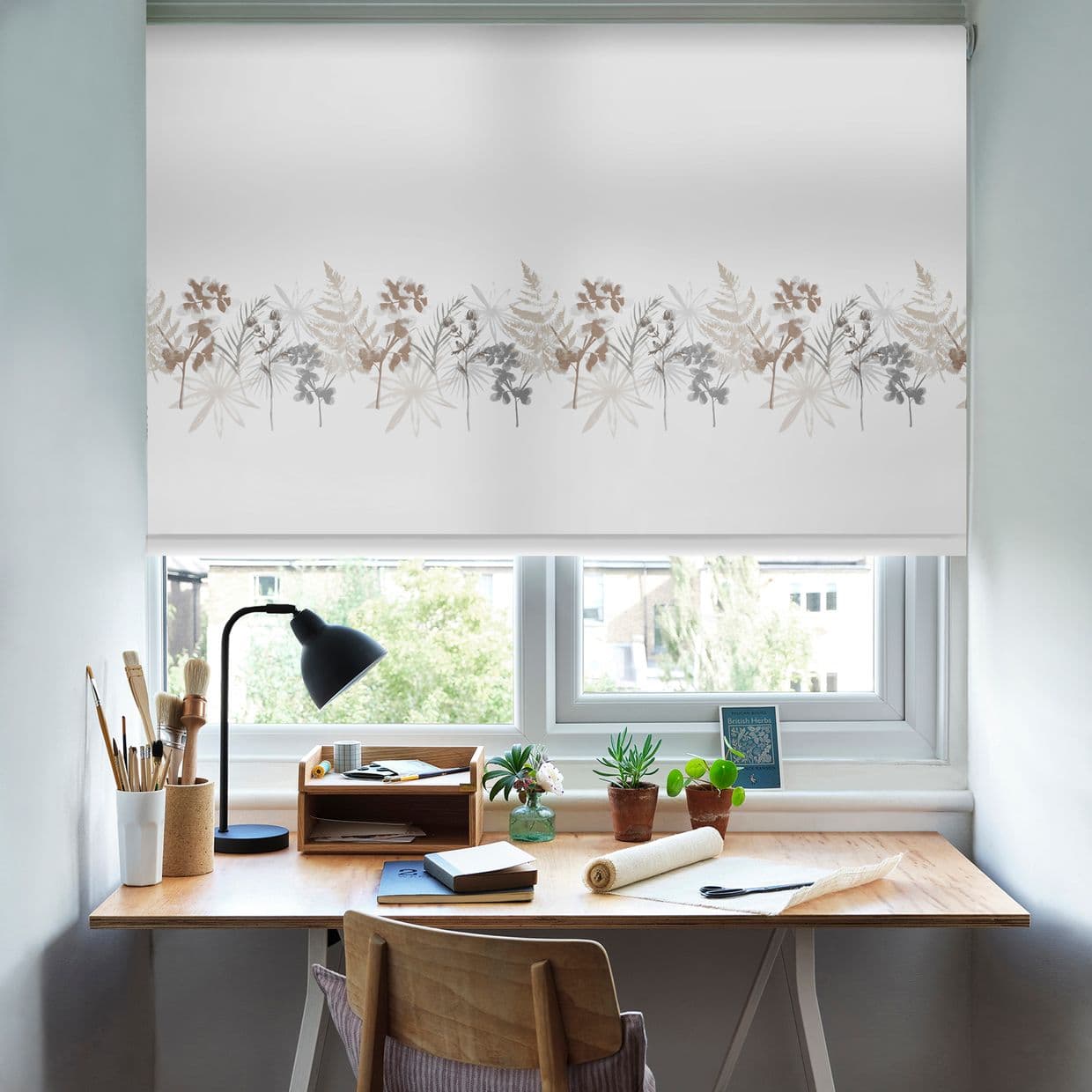 White roller blind with beige and gray botanical border, lowered across the window, filtering daylight. Above a wooden desk with lamp, potted plants, brushes and notebook in a bright home workspace.

British Herbs
