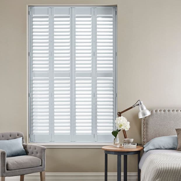 White full-height plantation shutters (four narrow panels) closed over the window, horizontal louvers slightly open and filtering bright daylight into a neutral-toned bedroom with armchair, bedside table, and lamp.
