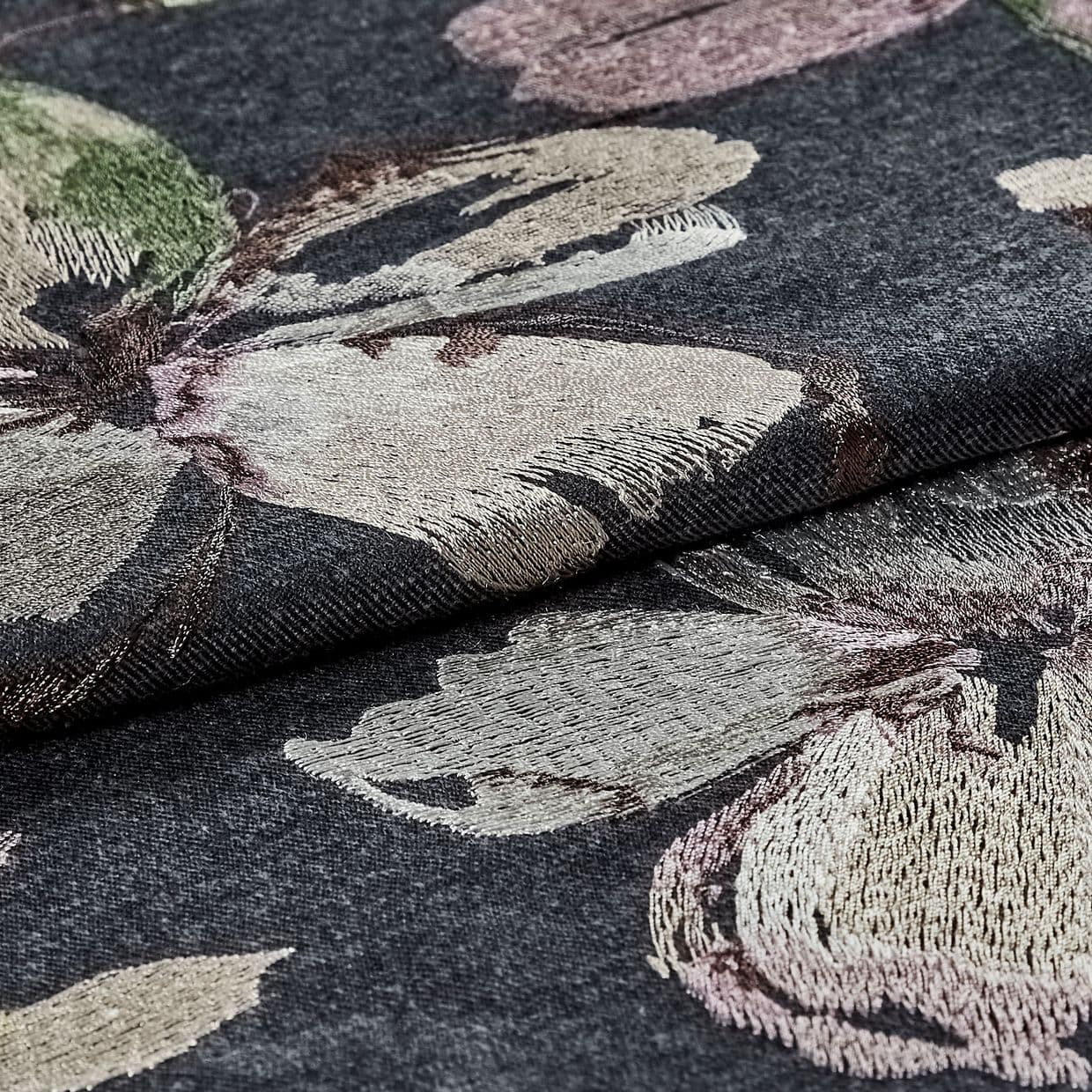 Dark grey jacquard fabric with large metallic-thread floral embroidery, folded and layered, showing textured, shimmering petals; close-up studio-lit view emphasizing weave, sheen, and textile detail.