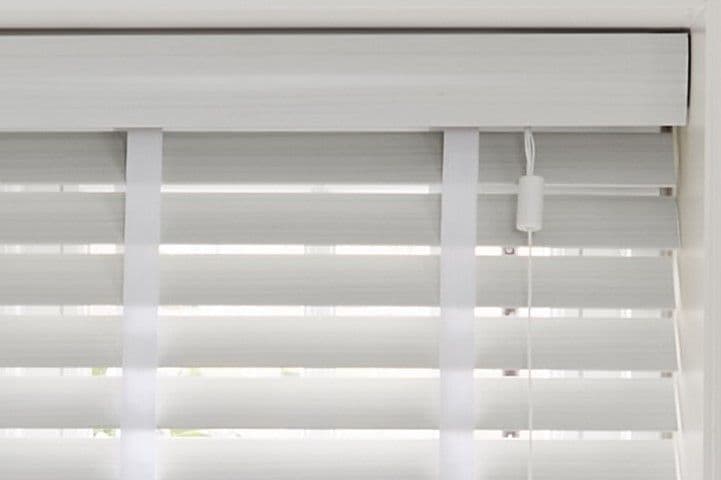 White horizontal blinds with wide slats and cloth ladder tapes, mounted inside the window frame; partially closed, filtering narrow bands of daylight into a bright, neutral interior.