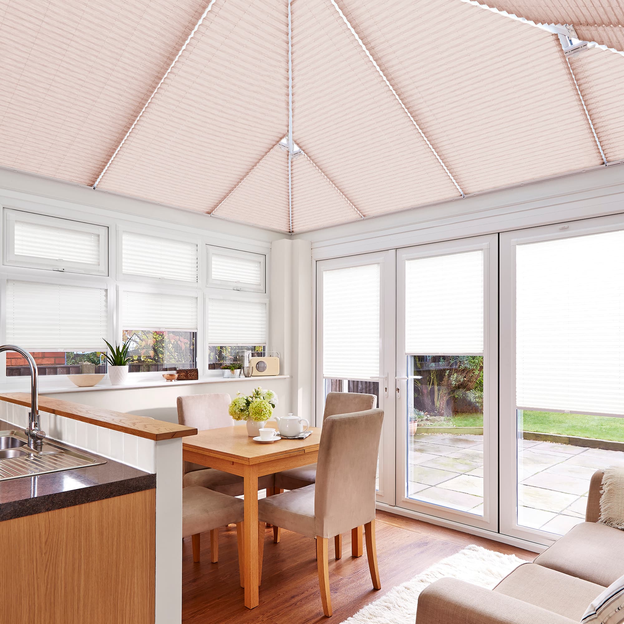 Pleated ceiling and window blinds, fully lowered, diffusing soft daylight; pale pink roof shades and white window blinds in a bright dining area with wooden table, upholstered chairs, and patio doors.