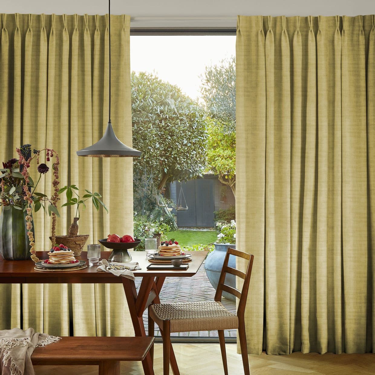 Floor-length pleated beige curtains flank a sliding glass door, partially drawn to reveal a sunlit garden; curtains filter warm light into a dining nook with wooden table, chairs, pendant lamp.