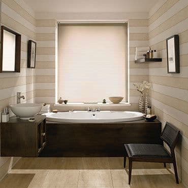 Cream roller blind fully lowered over central window, softly diffusing daylight; modern bathroom with built-in tub framed by dark wood paneling, striped beige walls, vessel sink, shelf, chair, and vases.
