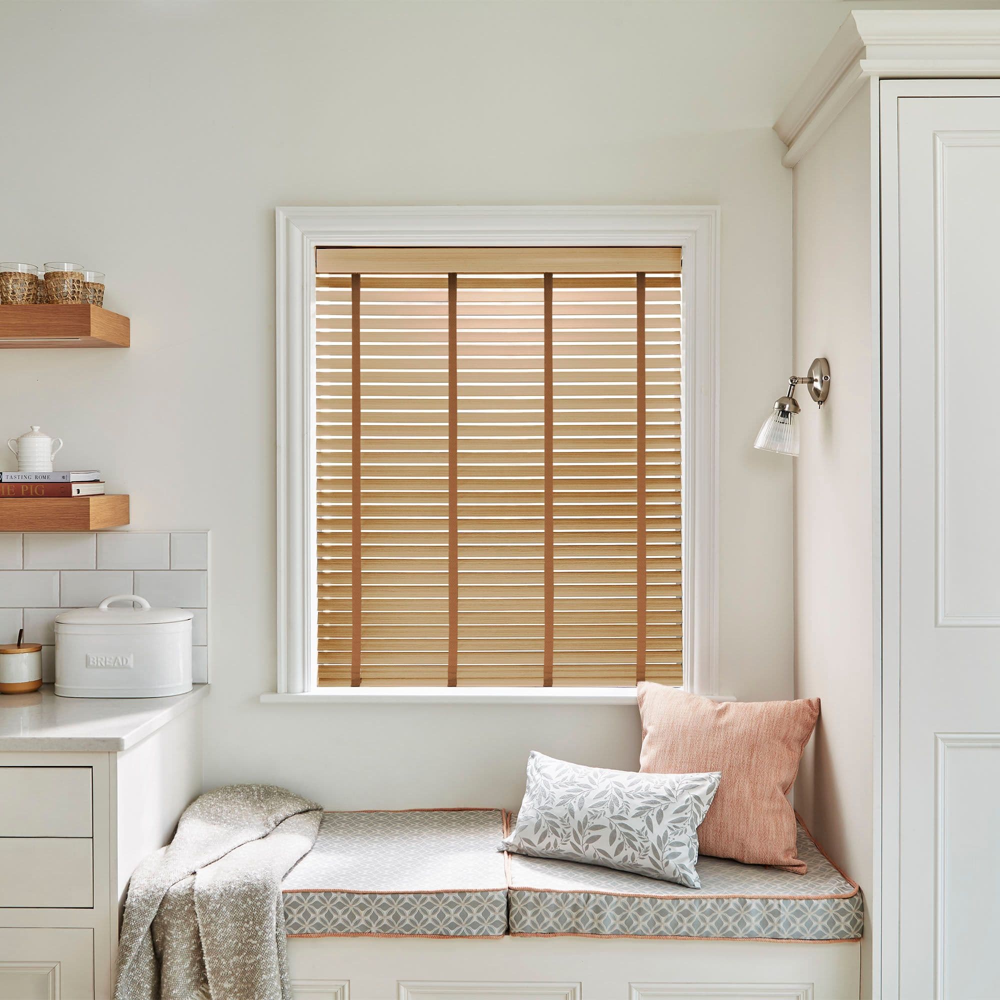 Wooden horizontal blinds with vertical tapes across the face, fully lowered and mostly closed, filtering warm daylight in a pale kitchen window-seat nook with cushions.

BREAD