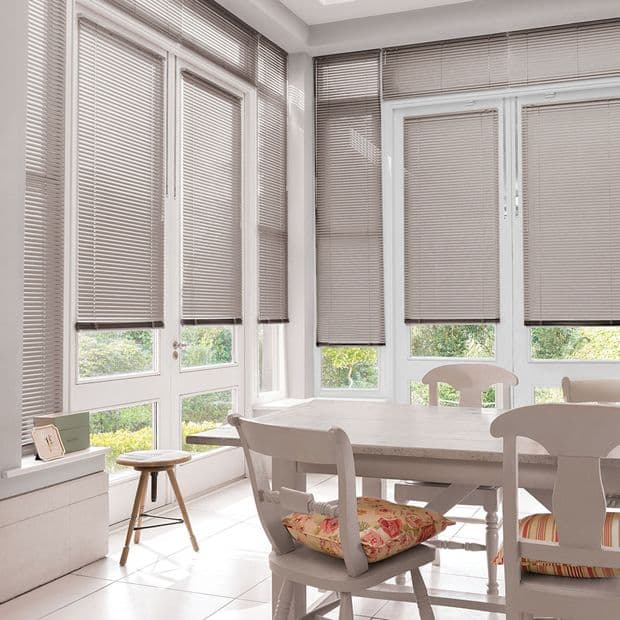Beige horizontal Venetian blinds covering tall corner French windows, lowered to mid-height and filtering daylight; a wooden dining table and chairs sit in a sunlit dining area overlooking a garden.