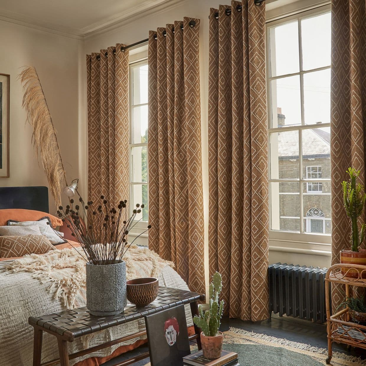 Floor-length brown geometric eyelet curtains hang closed along tall sash windows, filtering soft daylight into a cozy, bohemian bedroom with textured bedding, plants, and woven furnishings.