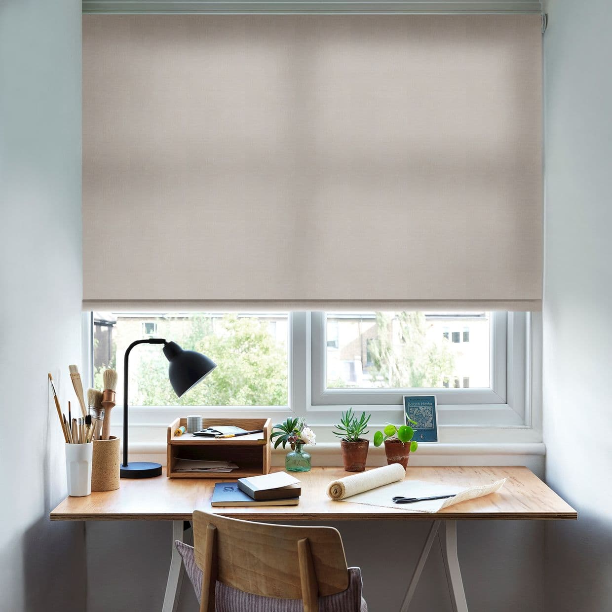 Beige roller blind, lowered nearly fully across the window, translucent fabric softening daylight; covering a small double-pane window above a wooden desk with lamp, potted plants and notebooks.