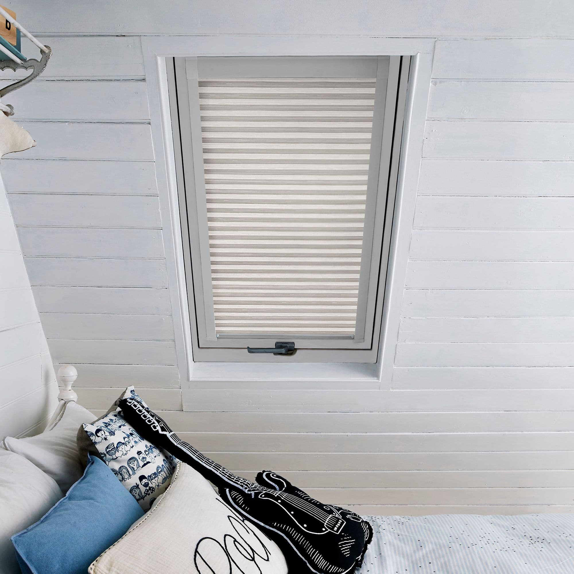 White pleated skylight blind, fully lowered across a recessed roof window, filtering soft daylight; set in white-painted tongue-and-groove attic ceiling above a bed with blue and patterned pillows.
