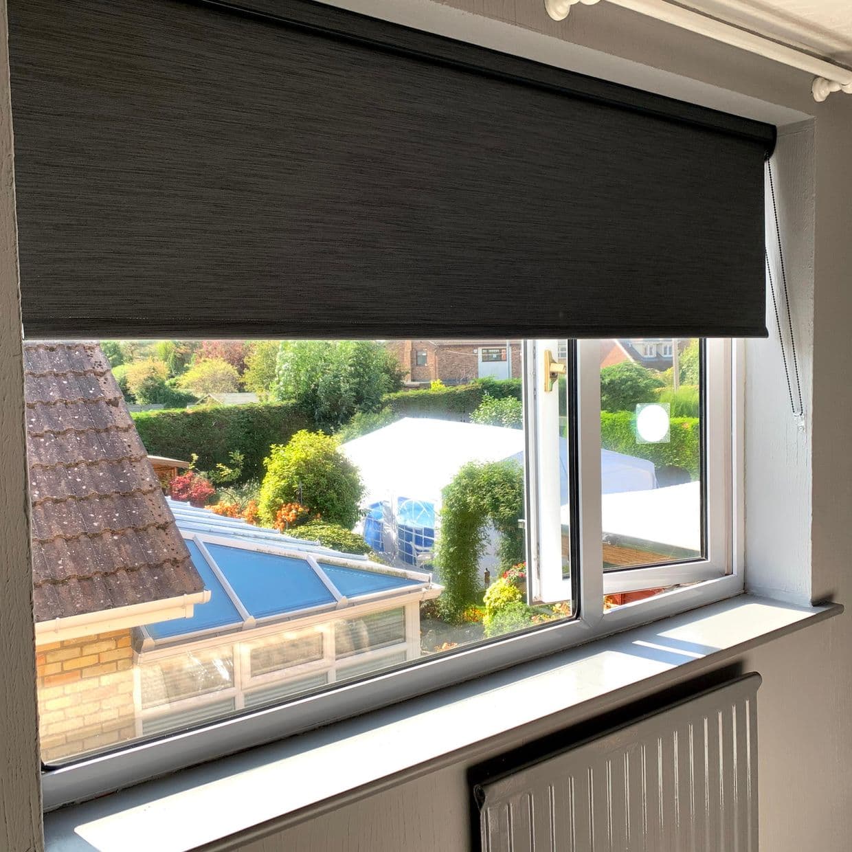 Dark grey roller blind lowered across the window with bead chain at right, partly filtering sunlight; view of sunlit suburban garden and conservatory roof, interior windowsill and radiator visible.