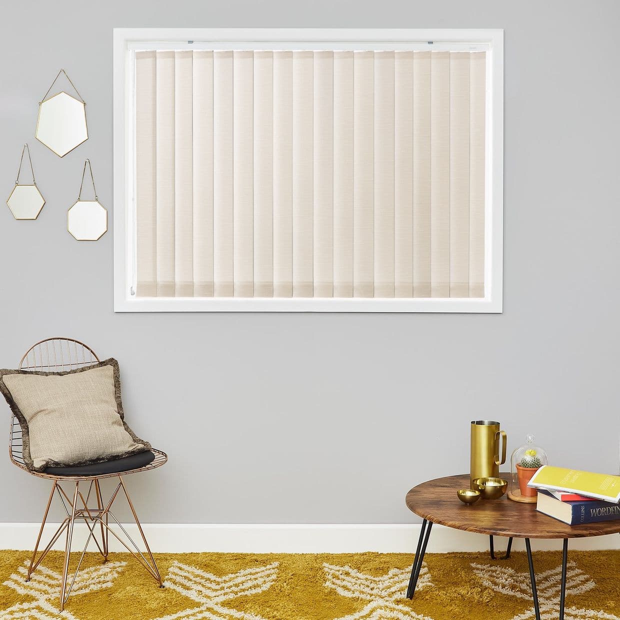 Beige vertical blinds, fully closed across a white-framed window. They filter soft daylight. Context: gray wall with three hanging hexagonal mirrors, wire chair with cushion, round wooden coffee table.