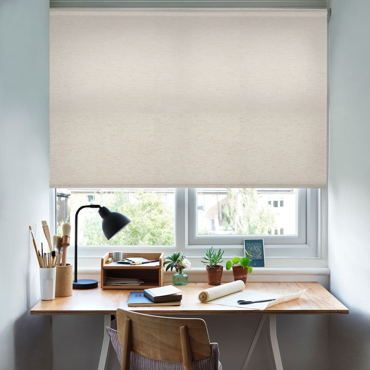 Beige fabric roller blind lowered across the window, filtering soft daylight; above a wooden desk with task lamp, brushes, notebooks, potted succulents, and a suburban view beyond the sill.