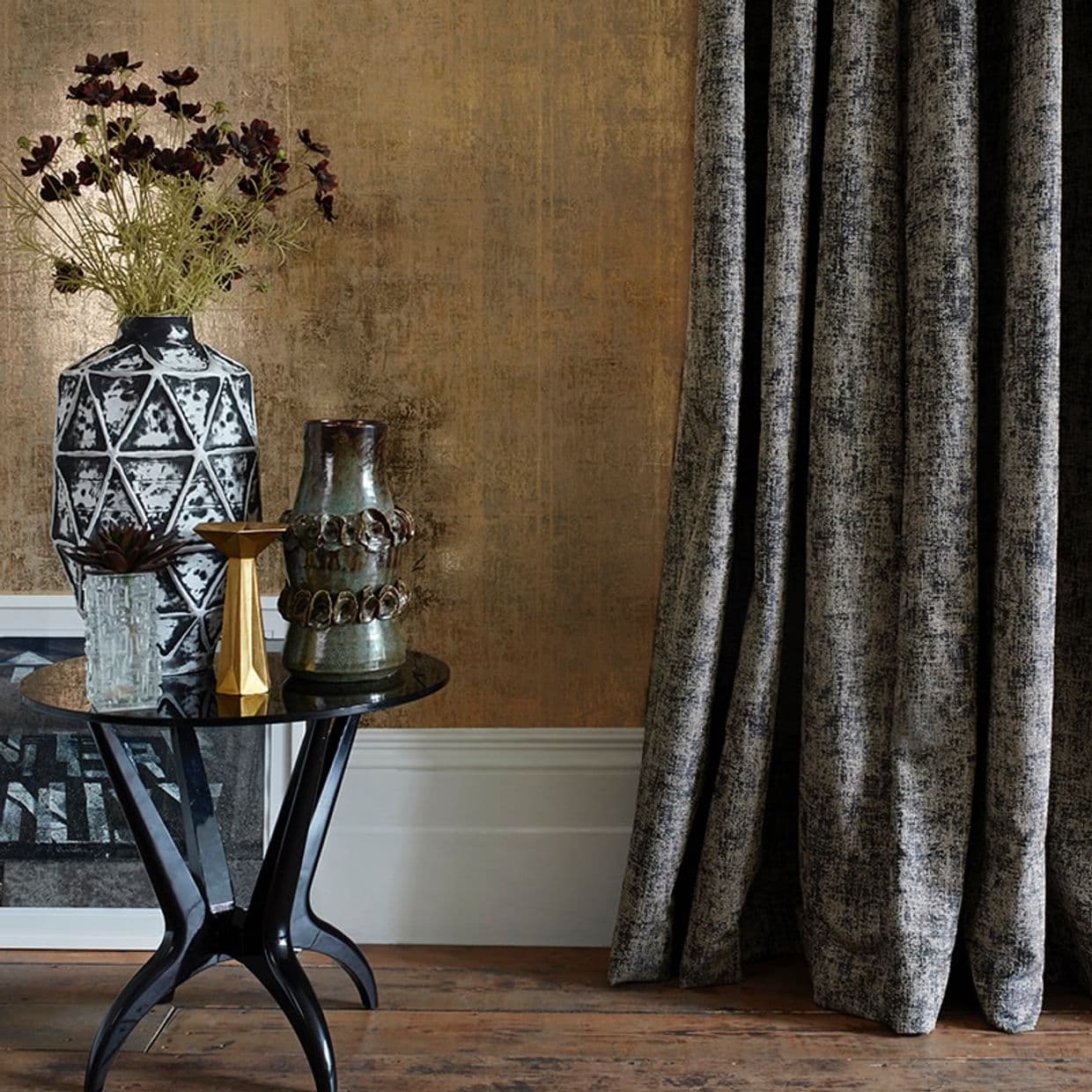 Heavy textured full-length grey curtain at right, hanging in pleats and pooling on a wooden floor; beside it a black side table with vases against a warm metallic-gold wall.