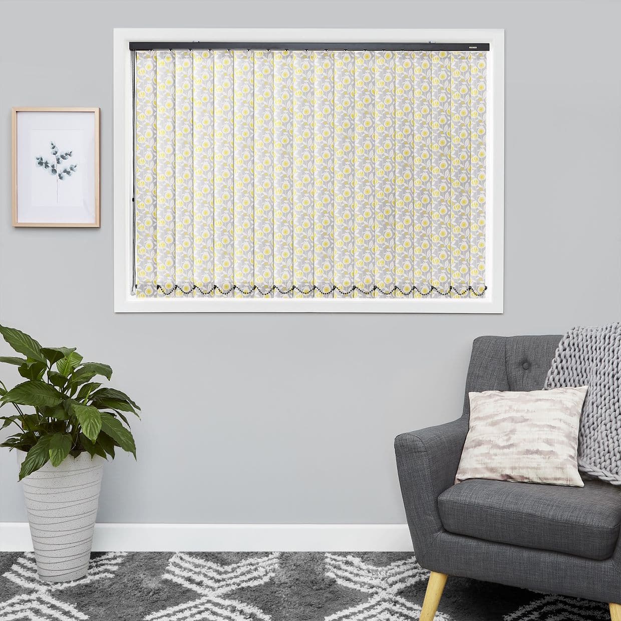 Fabric vertical blinds with yellow floral print, lowered and filtering daylight across a framed window; modern living room with gray walls, potted plant, patterned rug, and gray armchair.