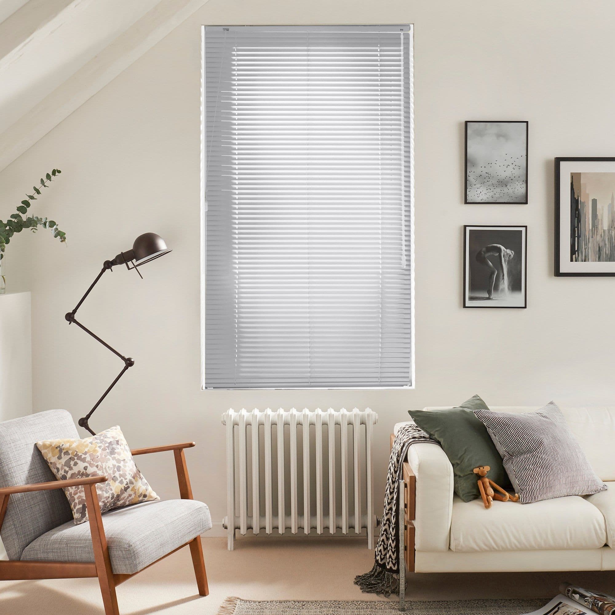 White horizontal Venetian blinds cover a tall window, closed and gently filtering daylight; in a neutral-toned living room with a radiator below, armchair, floor lamp, sofa and framed art.