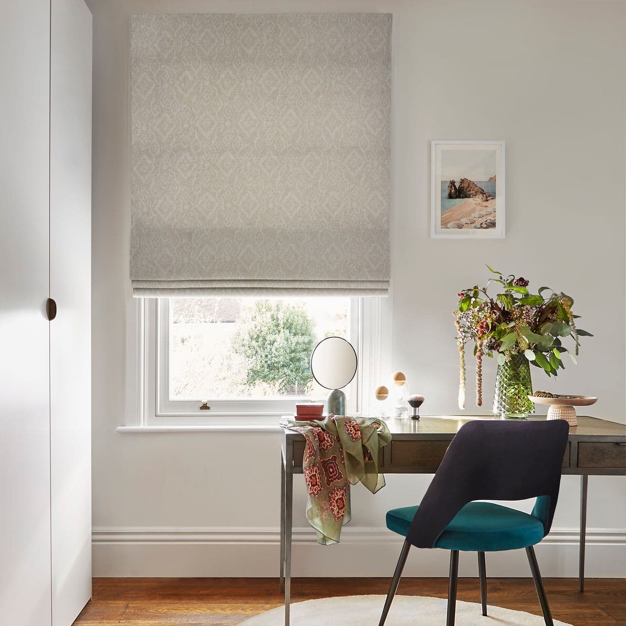 Roman patterned blind fully lowered with horizontal folds, filtering daylight. A desk and teal chair sit beneath in a light office/vanity, with mirror, vase of flowers and seaside print.