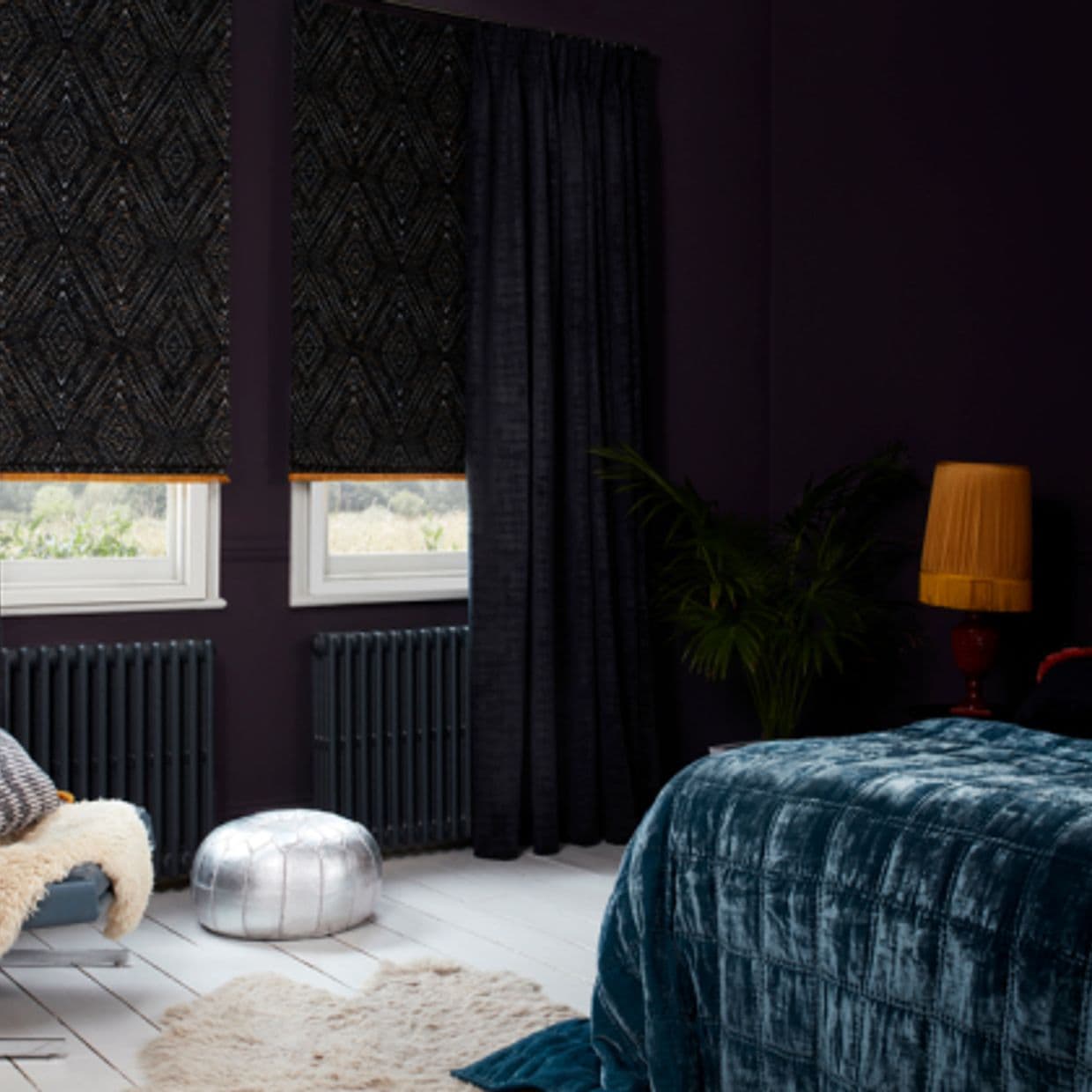 Dark patterned Roman shades with orange trim and heavy curtains cover twin windows; shades lowered, filtering light into a dim purple-walled bedroom with velvet bed, lamp and a potted plant.